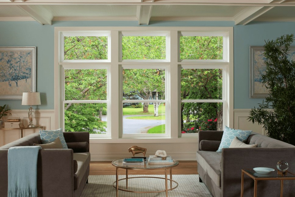 A spacious living room with a set of three vinyl windows looking out over a wooded front yard.
